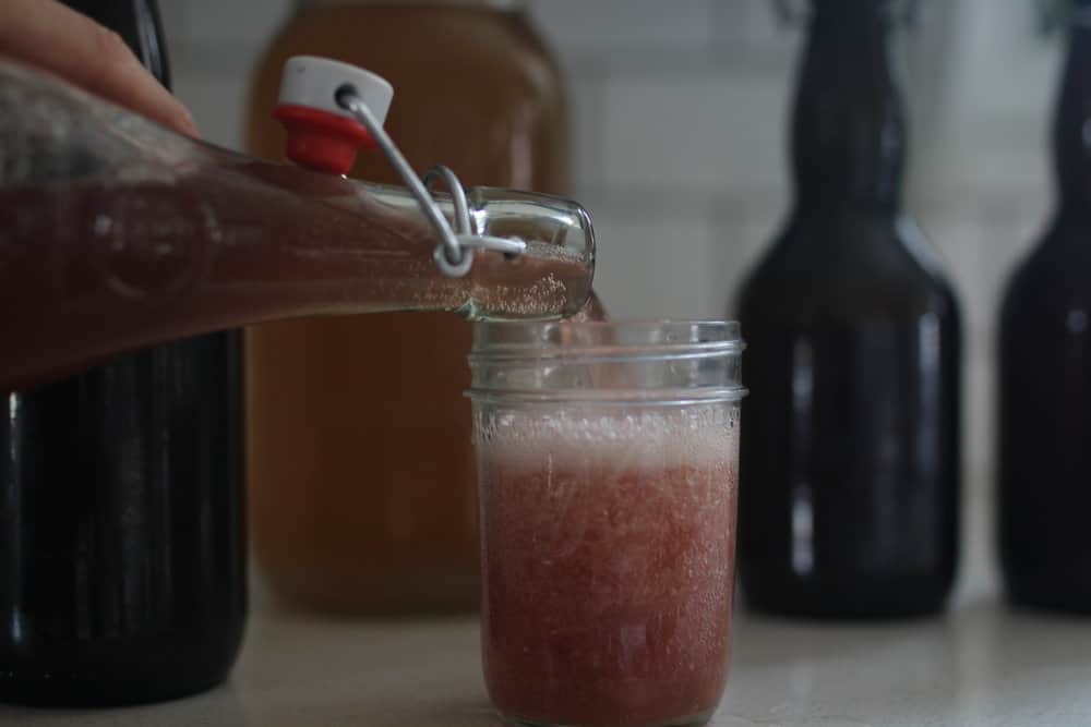 a glass bottle of water kefir soda is being poured into a glass mason jar with other bottles behind it
