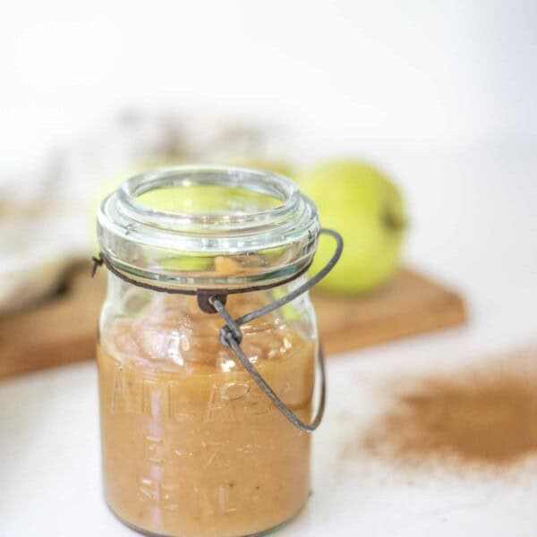 homemade applesauce in a vintage swing top jar on a white countertop with more apples in the background.