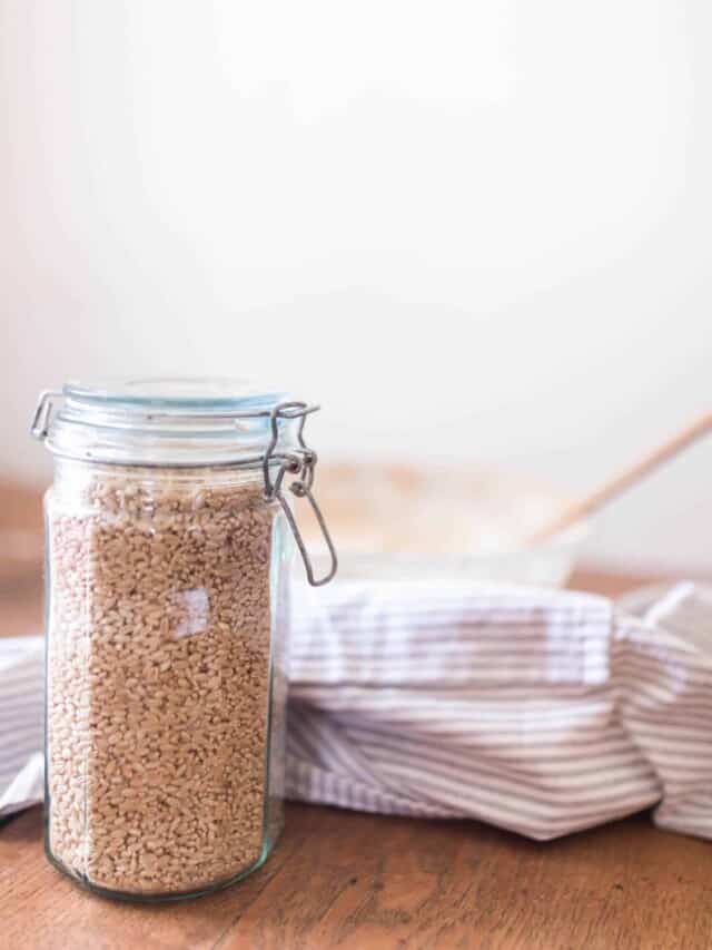 jar with a swing top lid full of wheat berries on a wood table with a bowl and linen stripped towel in the background