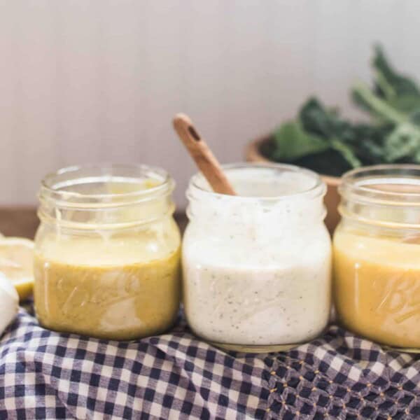 3 homemade dressings: honey mustard, ranch, and lemon vinaigrette dressing in mason jars on white and blue checked towels with a bowl of salad behind them.