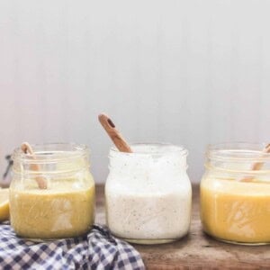 3 homemade salad dressings in glass jars with wooden spoons. On top a white and blue checked towel