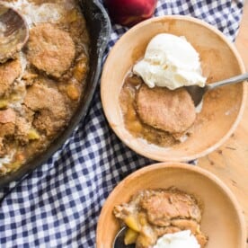 two bowls of peach cobbler in wooden bowls topped with ice cream on a blue and white checked towel and a cast iron skillet with remaining cobbler