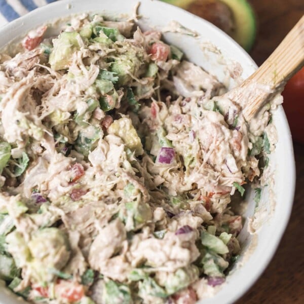 Southwest chicken salad in a white bowl with a wooden spoon sitting on a wood table with blue and white stripped towel and an avocado behind it