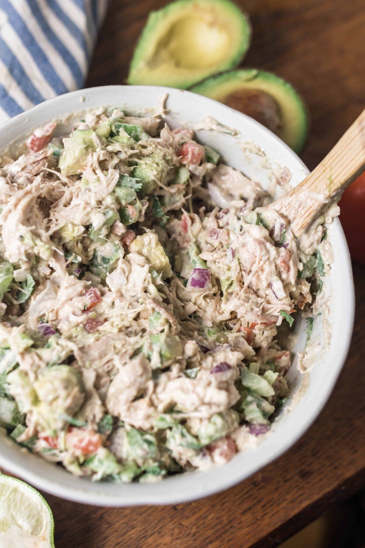 Southwest chicken salad in a white bowl with a wooden spoon sitting on a wood table with blue and white stripped towel and an avocado behind it.