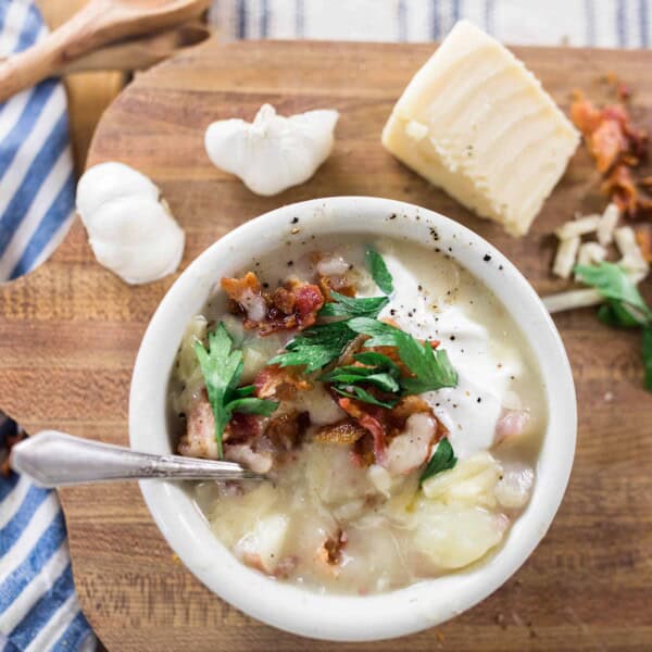 healthy potato sop in a white bowl topped with fresh herbs and bacon. Bowl of soup is on a wood cutting board topped with fresh ingredients and next to a blue and white stripped towel