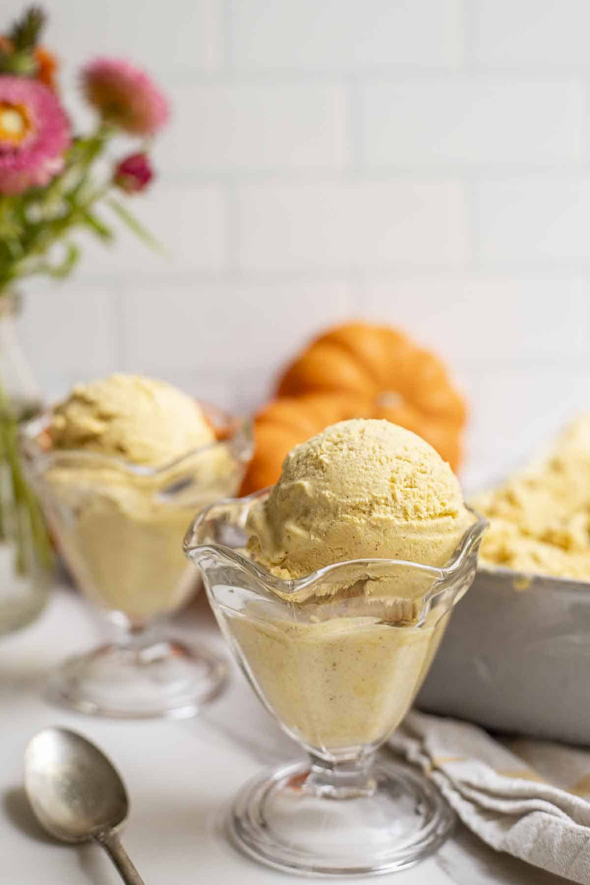 two ice cream cups filled with pumpkin ice cream. Pumpkins and flowers are in the background.