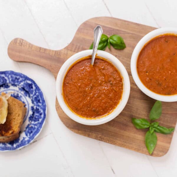 two bowls of roasted red pepper and tomato soup on a wood cutting board with basil. Grilled cheese on a blue and white plate is to the left