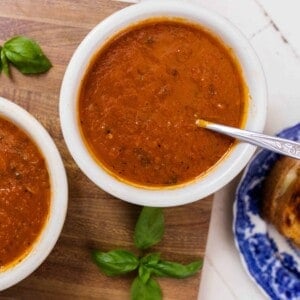 two bowls filled with of roasted red pepper and tomato soup on top a cutting board with a blue plate of grilled cheese