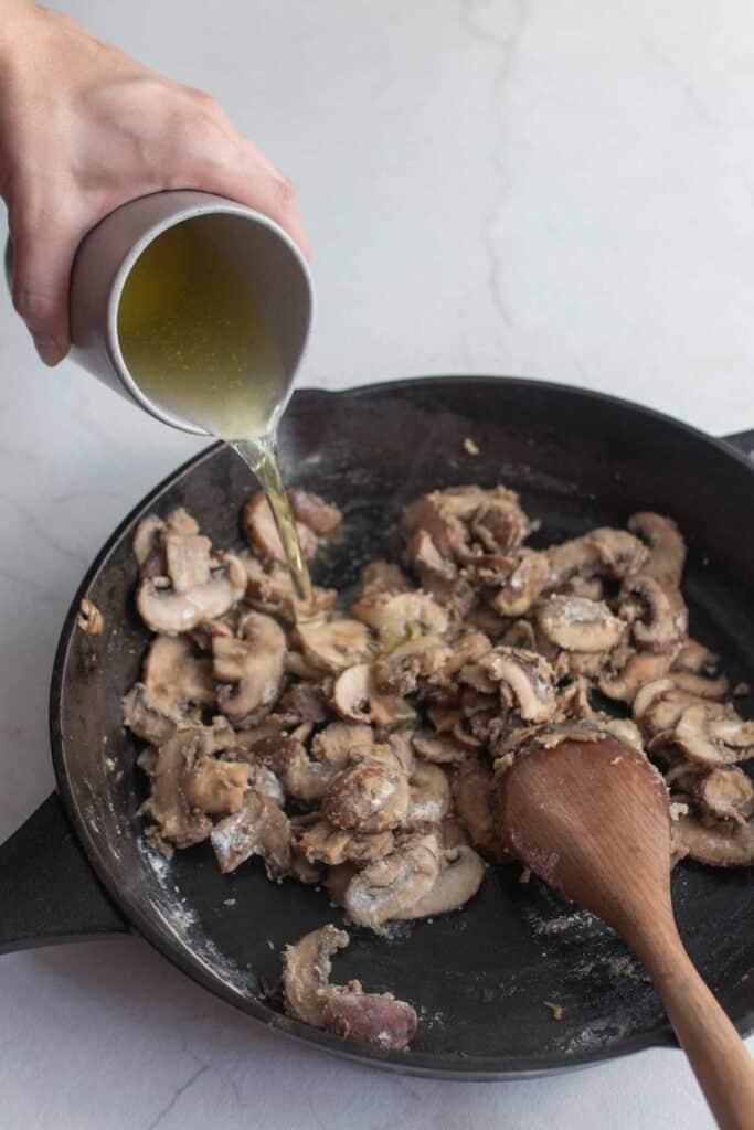 A hand pouring broth into a cast iron skillet filled with sauted sliced mushrooms.