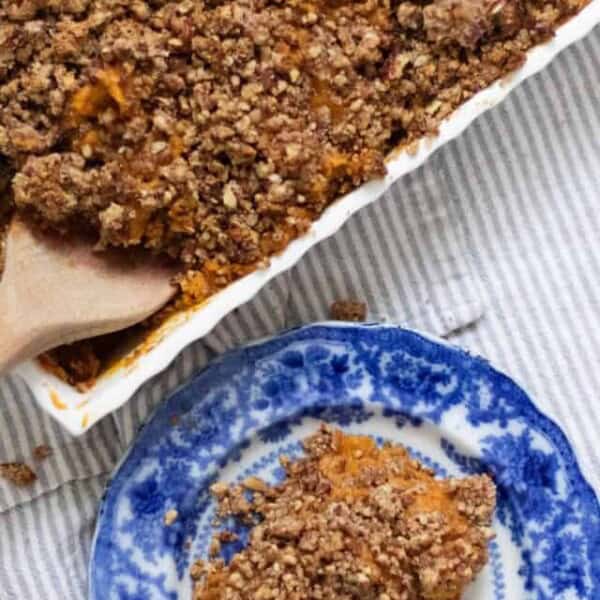 casserole dish of healthy sweet potato casserole with a pecan crumble with a wooden spoon in the dish. A antique blue plate sits to the right with sweet potato casserole on it.