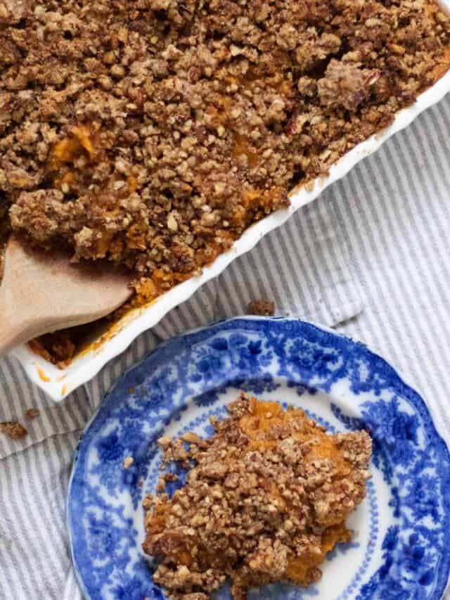 casserole dish of healthy sweet potato casserole with a pecan crumble with a wooden spoon in the dish. A antique blue plate sits to the right with sweet potato casserole on it.