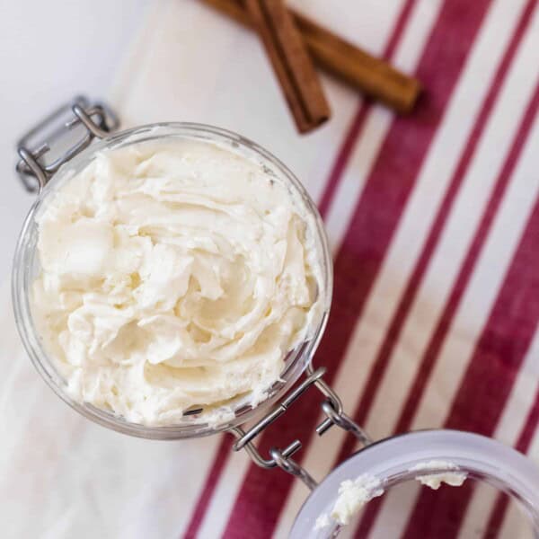 overhead shot of gingerbread whipped body butter in a glass jar on a red and cream stripped towel next to cinnamon sticks