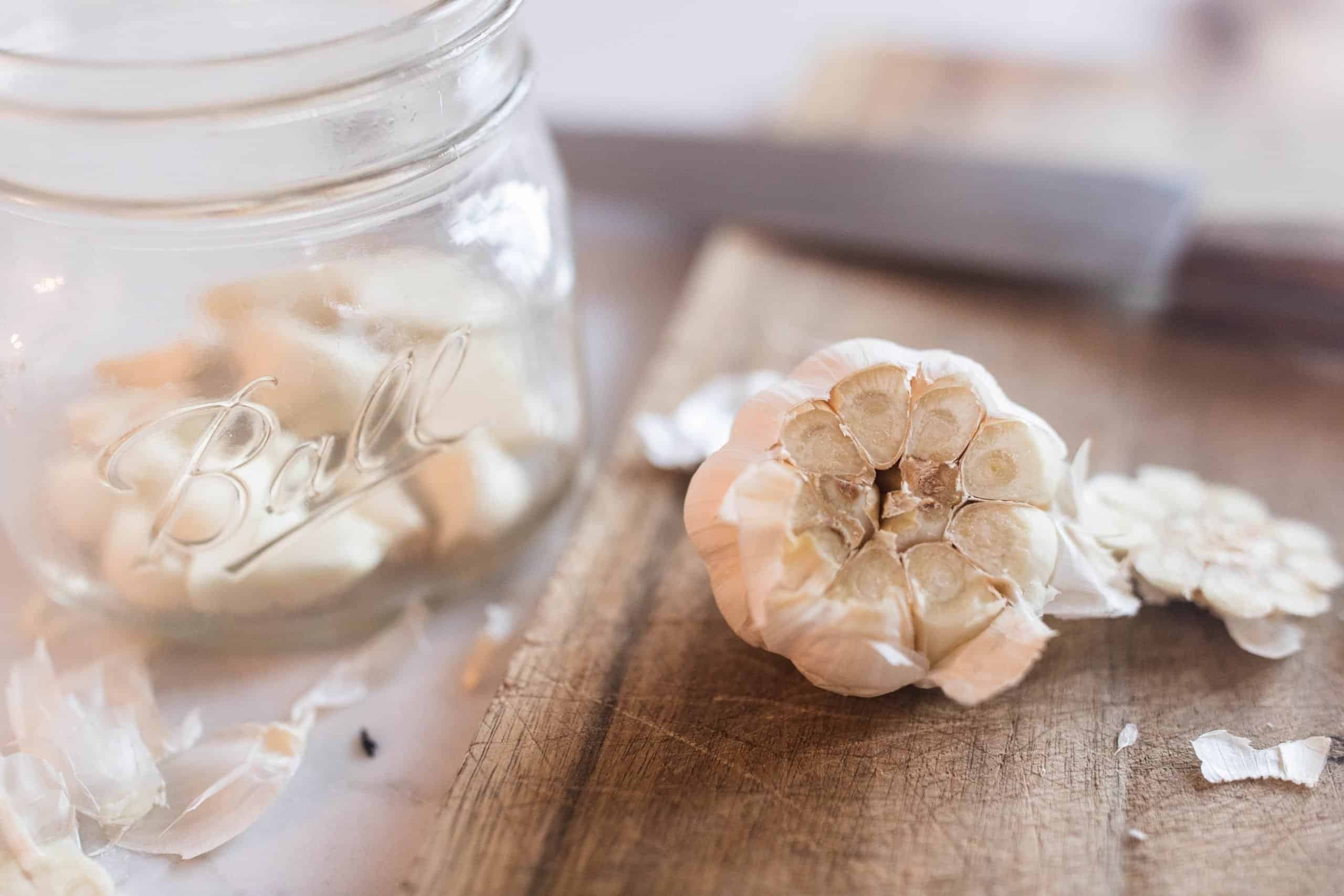 head of garlic with the top cut off with a jar of peeled garlic cloves in a jar to the left.