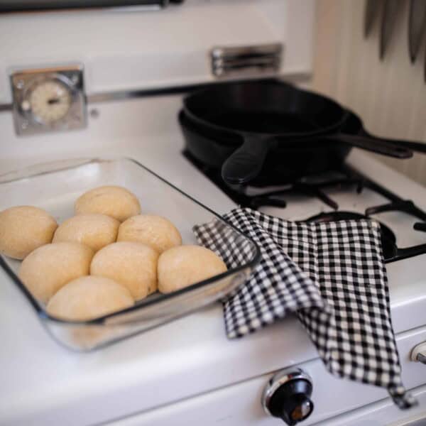 sourdough rolls in a baking dish on top a antique oven with a blue and white plaid towel to the right and cast iron skillet in the background.