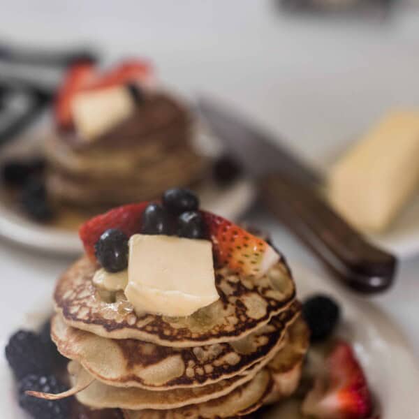 two stacks of buckwheat pancakes topped with fresh fruit and butter on white places