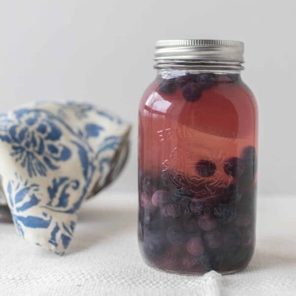 Fermented blueberries in a mason jar with a silver bowl covered with a blue and white damask printed towel in the background