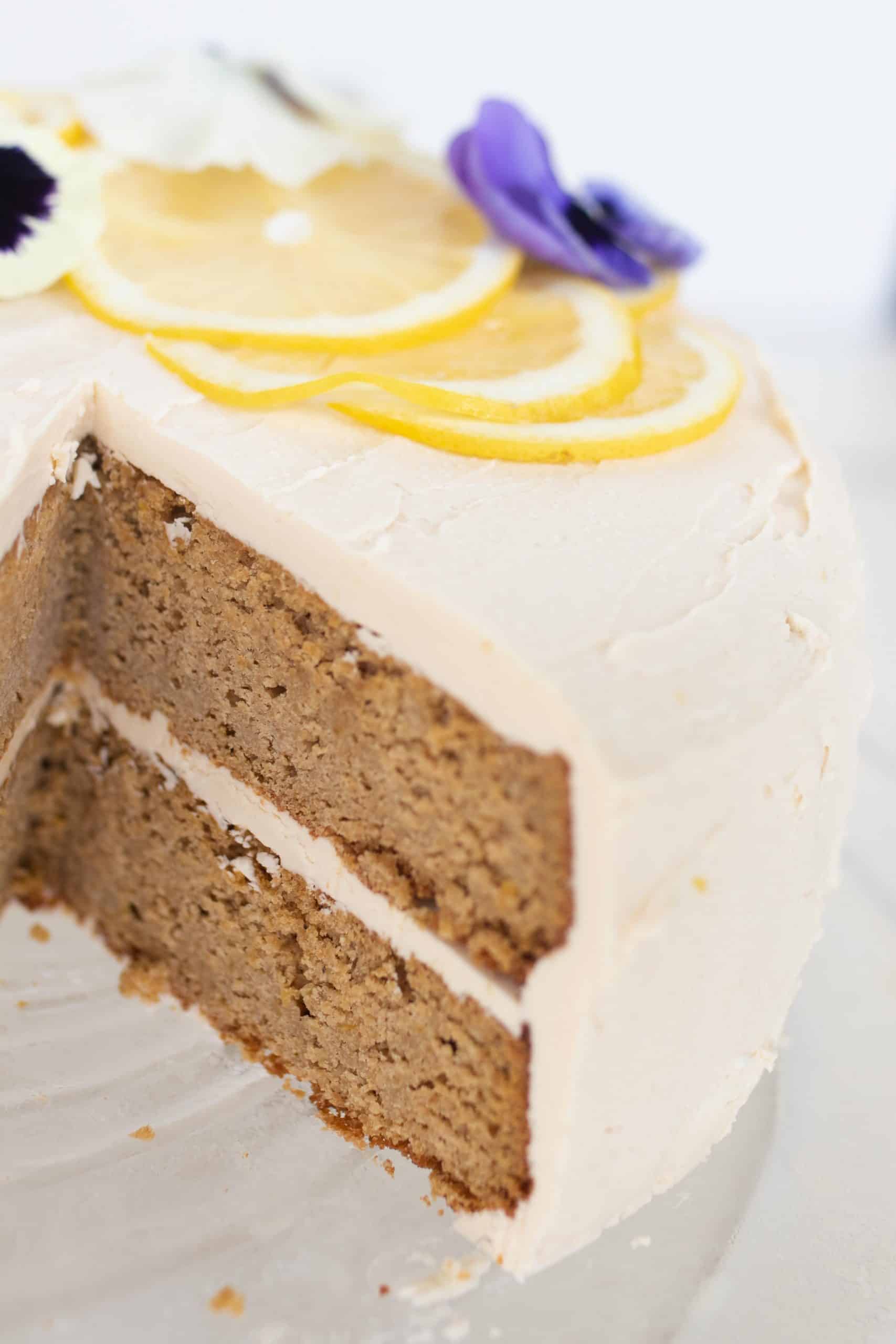 close up picture of a two tiered gluten free lemon cake lemon icing with a slice taken out. The cake is topped with lemon slices and edible flower.