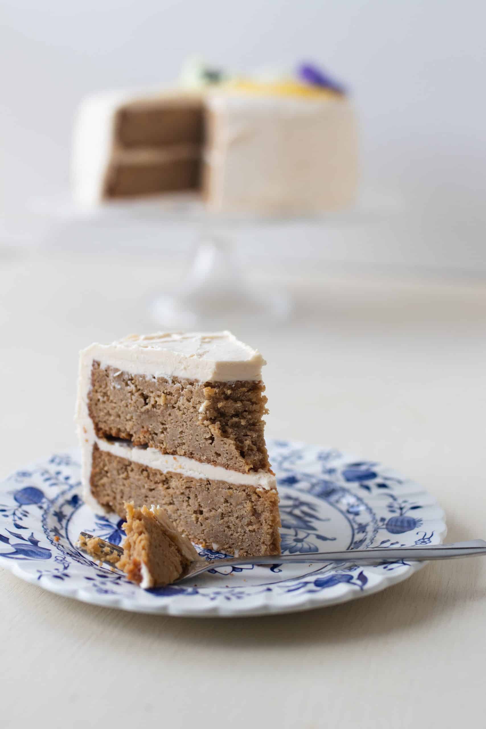 a slice of two tiered gluten free lemon cake with lemon icing on a blue and white plate with a fork. The remaining cake sits on a cake stand in the background