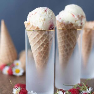 cones filled with strawberry cheesecake ice cream in cones in glasses on a wood table with strawberries and flowers spread across the table.