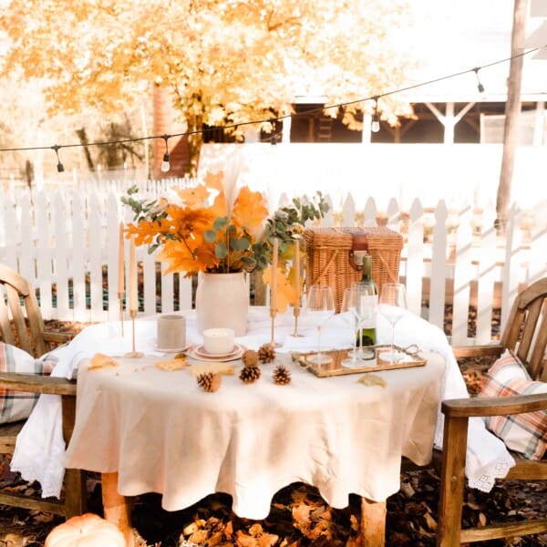 wooden table with tan linen table cloth with a crock of dried leaves and eucalyptus with a picnic basket with a barn with a picket fence in the background