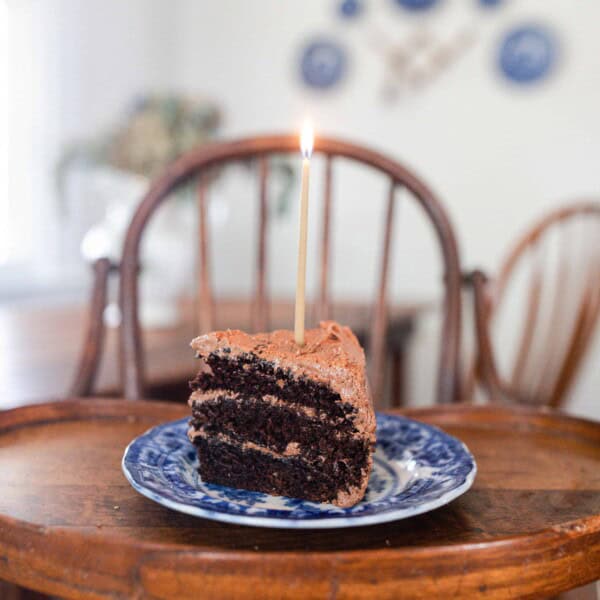 slice of three layer sourdough chocolate cake with chocolate frosting on a blue and white antique plate with a candle on a antique wooden high chair