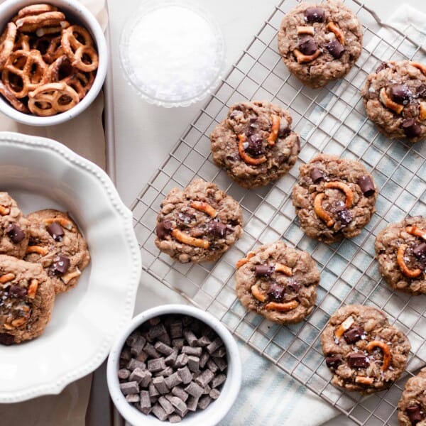 overhead photo of a cooling rack of chocolate oatmeal lactation cookies with a plate of cookies to the left and two white dishes of pretzels and chunks