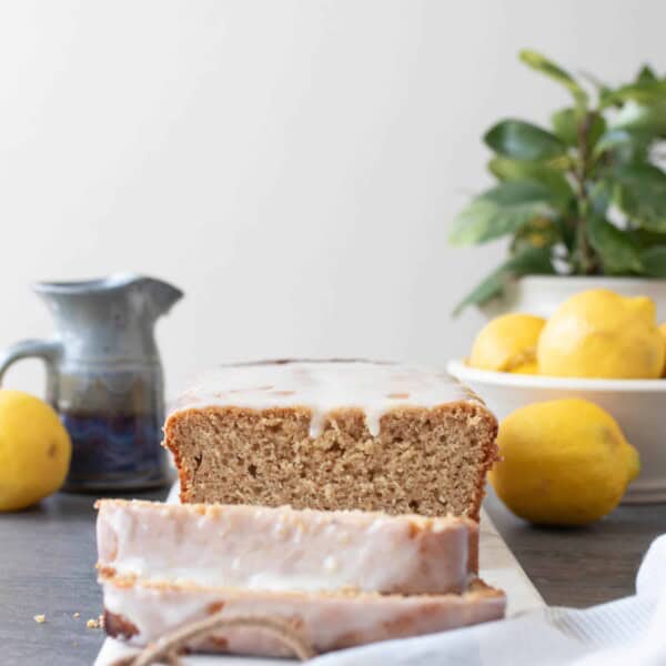 sourdough lemon pound cake with a lemon glaze sliced on a marble cutting board. A small gray pitcher, lemons, and a potted plant in the background