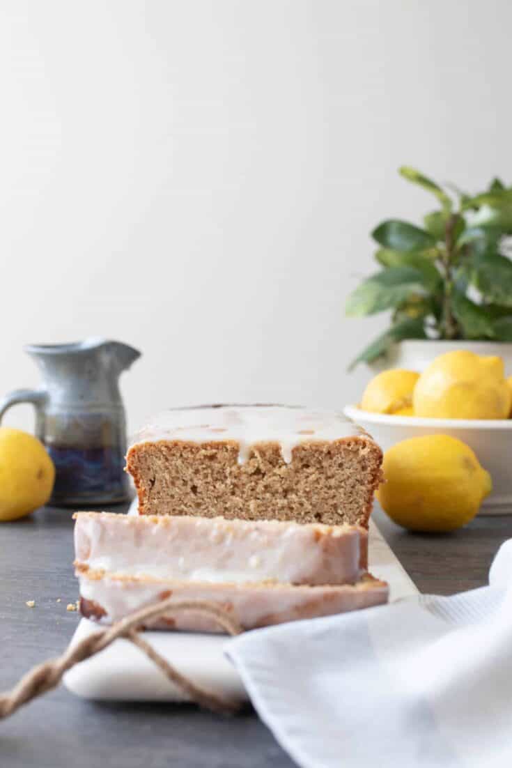 sourdough lemon pound cake with a lemon glaze sliced on a marble cutting board. A small gray pitcher, lemons, and a potted plant in the background