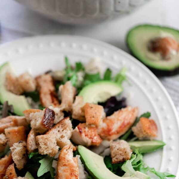 a white plate with a greens, avocados, and sourdough croutons with a bowl of croutons and a half of avocado in the background