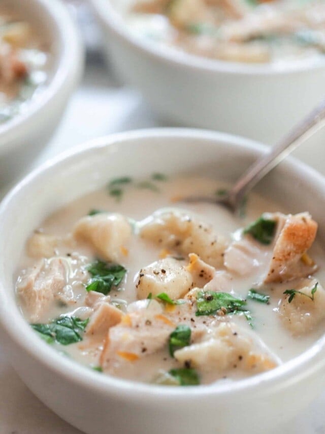 creamy gnocchi soup with chicken and herbs in a white bowl with a spoon in the soup. More bowls of soup in the background