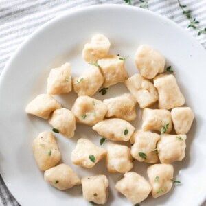 overhead photo of a white bowl full of homemade gnocchi with fresh herbs sprinkled on top. a white and blue stripped towel topped with fresh thyme sits behind the bowl.