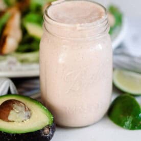 Mexican ranch dressing in a mason jar on a white counter with sliced avocados, limes around the jar and a salad in the background