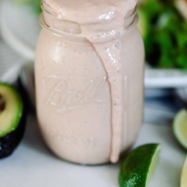 Mexican ranch dressing in a mason jar wit the dressing spilling over the top. Sliced limes and avocados lay on the counter around the jar of ranch