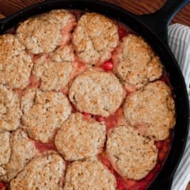 overhead photo of sourdough cherry cobbler in a cast iron skillet on top of a blue and white stripped towel laying on a wooden table