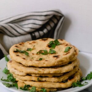 einkorn flat bread stacked with herbs sprinkled over on a white plate with a gray and black stripped tea towel in the background