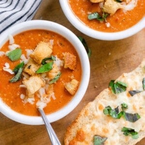 two bowls of tomato soup from scratch with fresh tomatoes topped with fresh basil, croutons, and cheese on a wood cutting board with a loaf of cheesy bread and a white and black stripped towel to the left