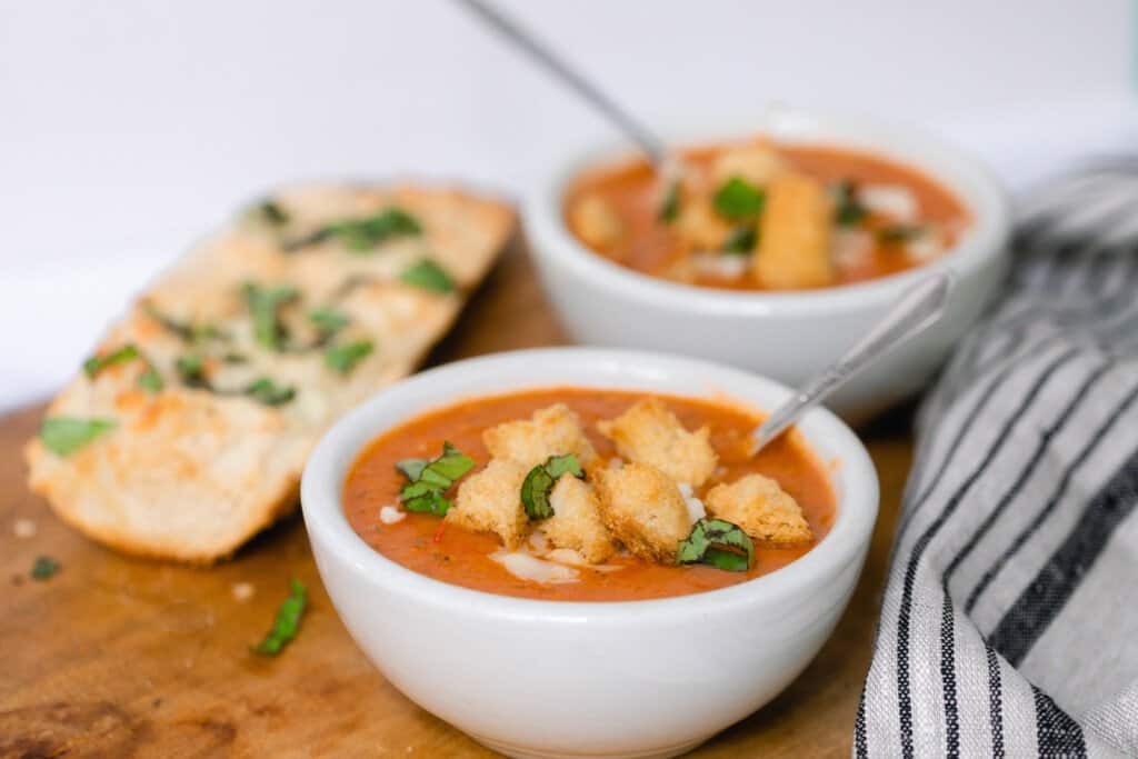 two bowls of tomato soup topped with fresh herbs and croutons on a wooden cutting board with some garlic herb bread behind the bowls