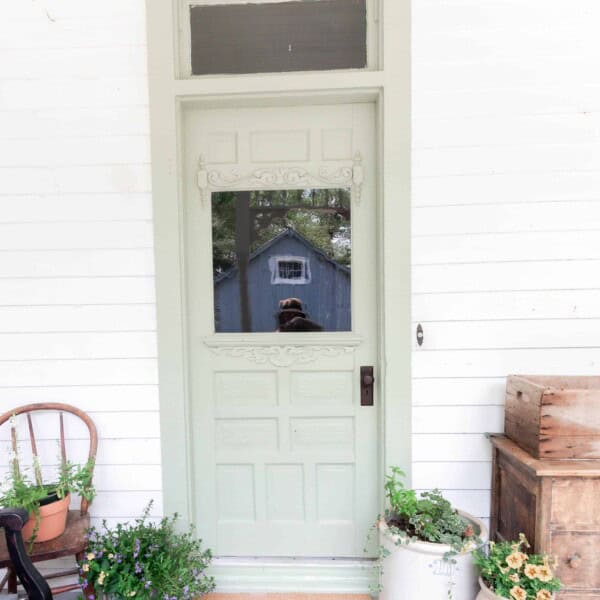 sage green farmhouse front door with a DIY Cricut doormat. Crocks with flowers sit to the right and a antique wooden chair with flowers in a pot on the right of the door