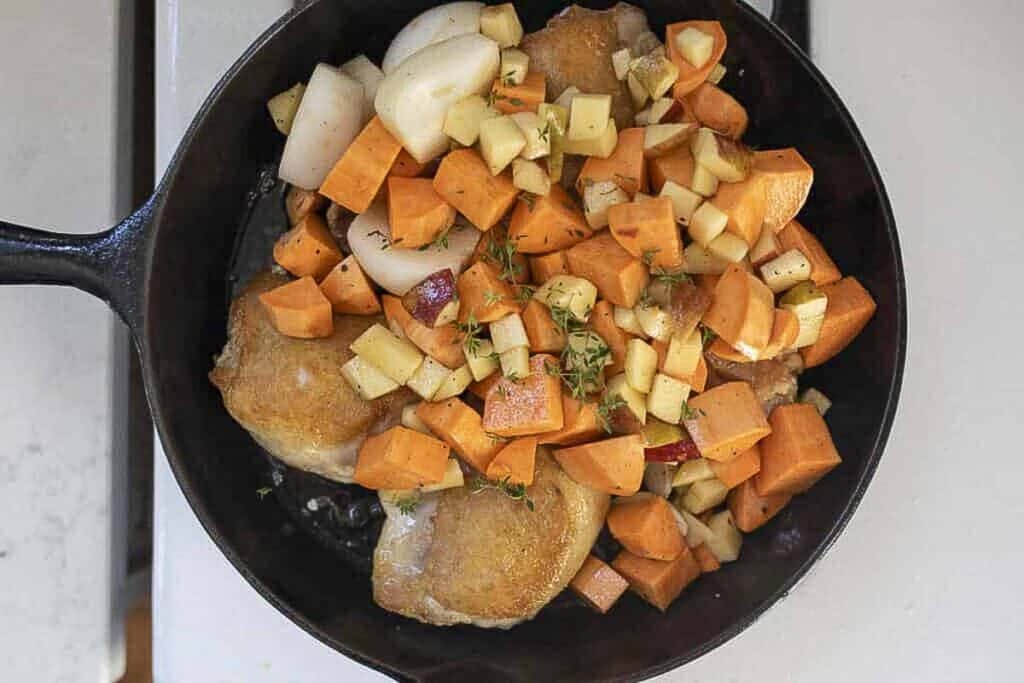 adding sweet potatoes, onions, and apples to the chicken in a skillet.