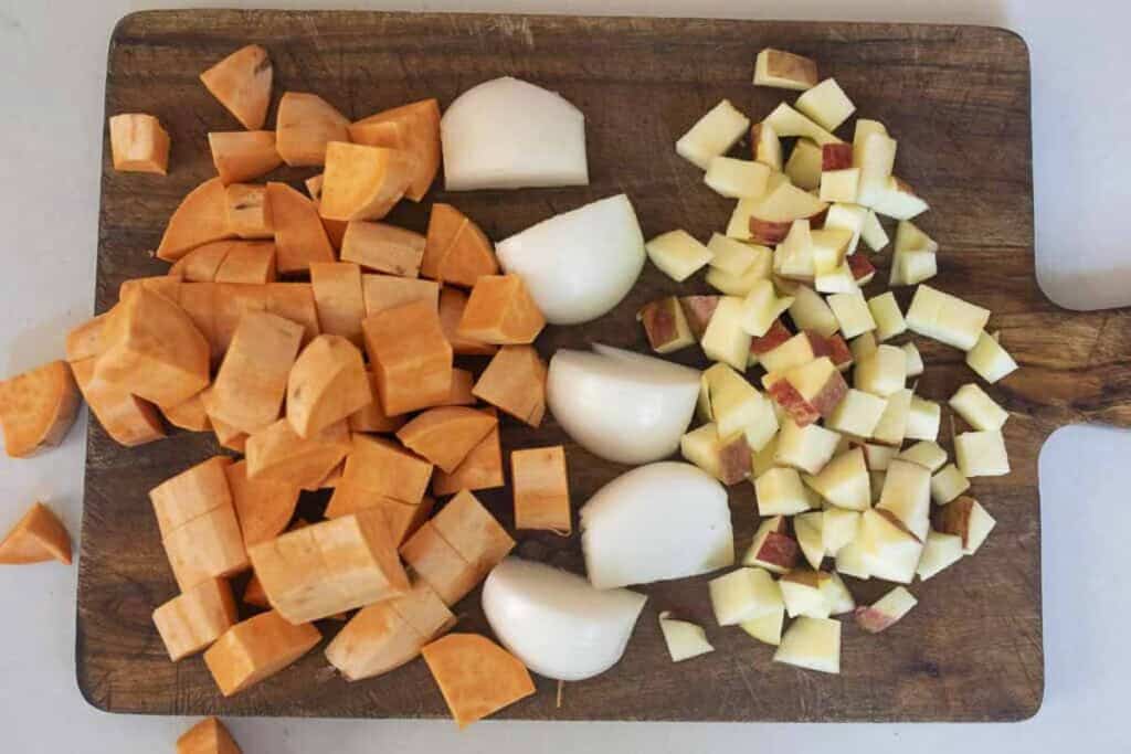 apples, sweet potatoes, and an onion chopped on a wood cutting board.