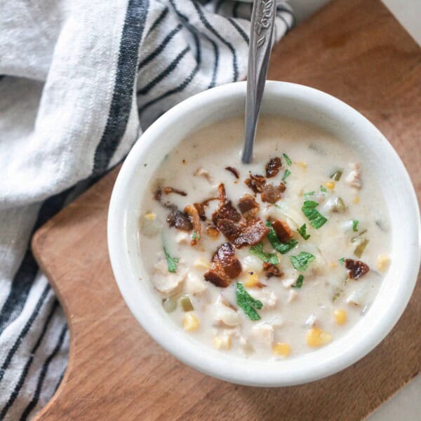 overhead photo of chicken corn chowder topped with bacon and fresh herbs on a wood cutting board on a black and white stripped towel