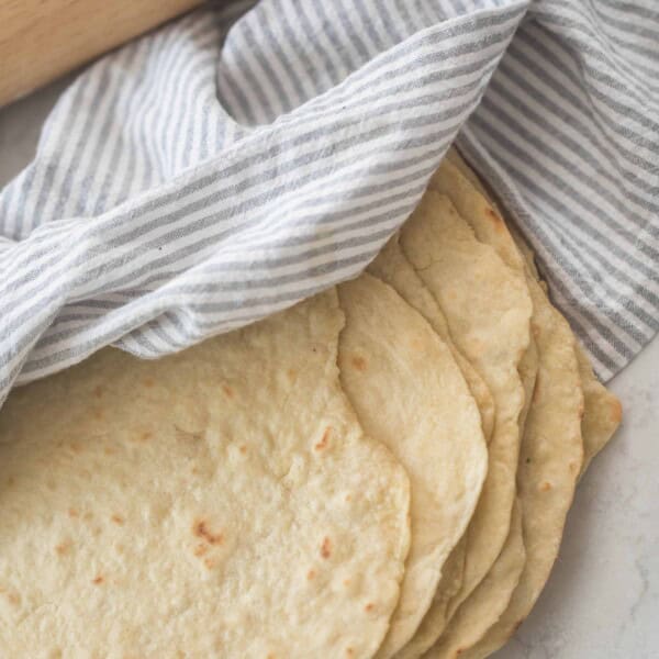 homemade einkorn tortillas stacked on a white quartz countertop. a gray and white towel is draped over the tortillas and a rolling pin is in the background