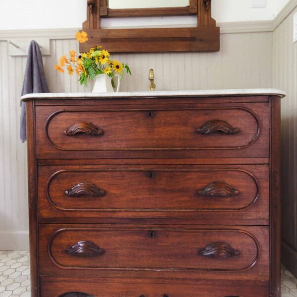 stained wooden antique dresser turned into a bathroom vanity with a marble top. A antique mirror hangs over the vanity.