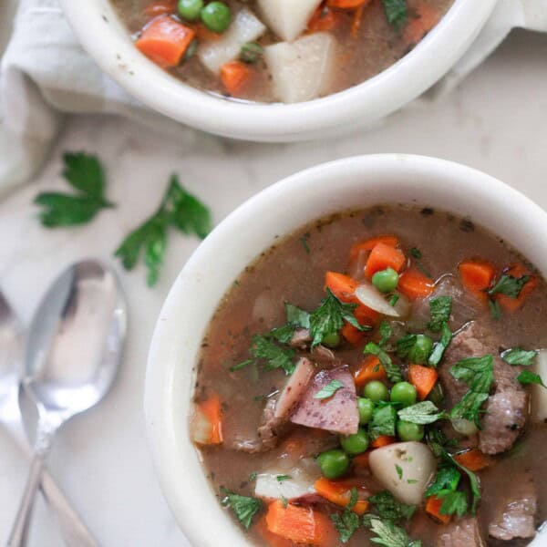 overhead photo of two bowls of beef stew on a white countertop with two spoons to the left of one bowl and a napkin in the background