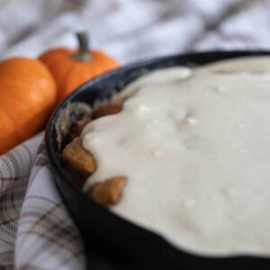 sourdough pumpkin cinnamon rolls covered in cream cheese frosting in a cast iron skillet on a brown plaid napkin with two small pumpkins to the back left