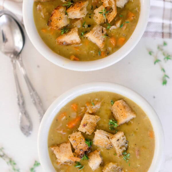 overhead photo of two bowls of homemade split pea soup topped with thyme and sourdough croutons on a white countertop and two spoons to the left and a plaid towel in the top right corner.