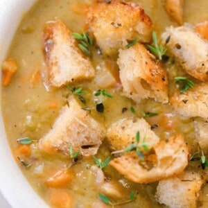 close up overhead photo of split pea soup topped with sourdough croutons with chunks of carrots and fresh thyme in a white bowl