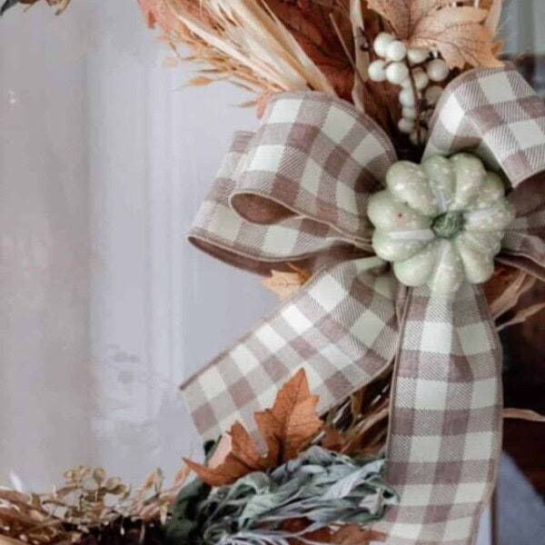 a fall wreath made with faux leaves, wheat, dried sage and tied with a tan and white checked ribbon with a light green pumpkin in the middle.