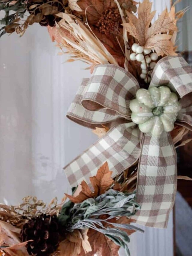 a fall wreath made with faux leaves, wheat, dried sage and tied with a tan and white checked ribbon with a light green pumpkin in the middle.