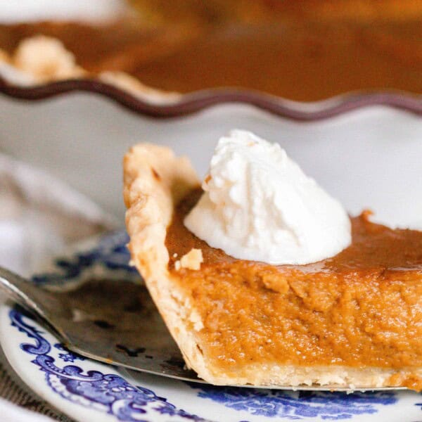 slice of pumpkin pie made from scratch with a dollop of whipped cream on a antique blue and white plate. The rest of the pie is in the background